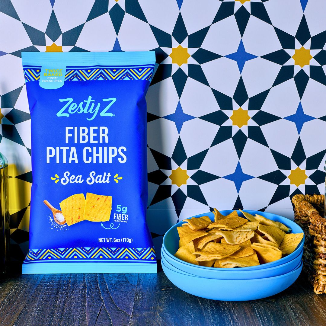 A bag of ZestyZ Fiber Pita Chips Sea Salt stands on a dark wooden surface next to a blue bowl filled with pita chips, set against a geometric navy, white, and yellow patterned tile wall.