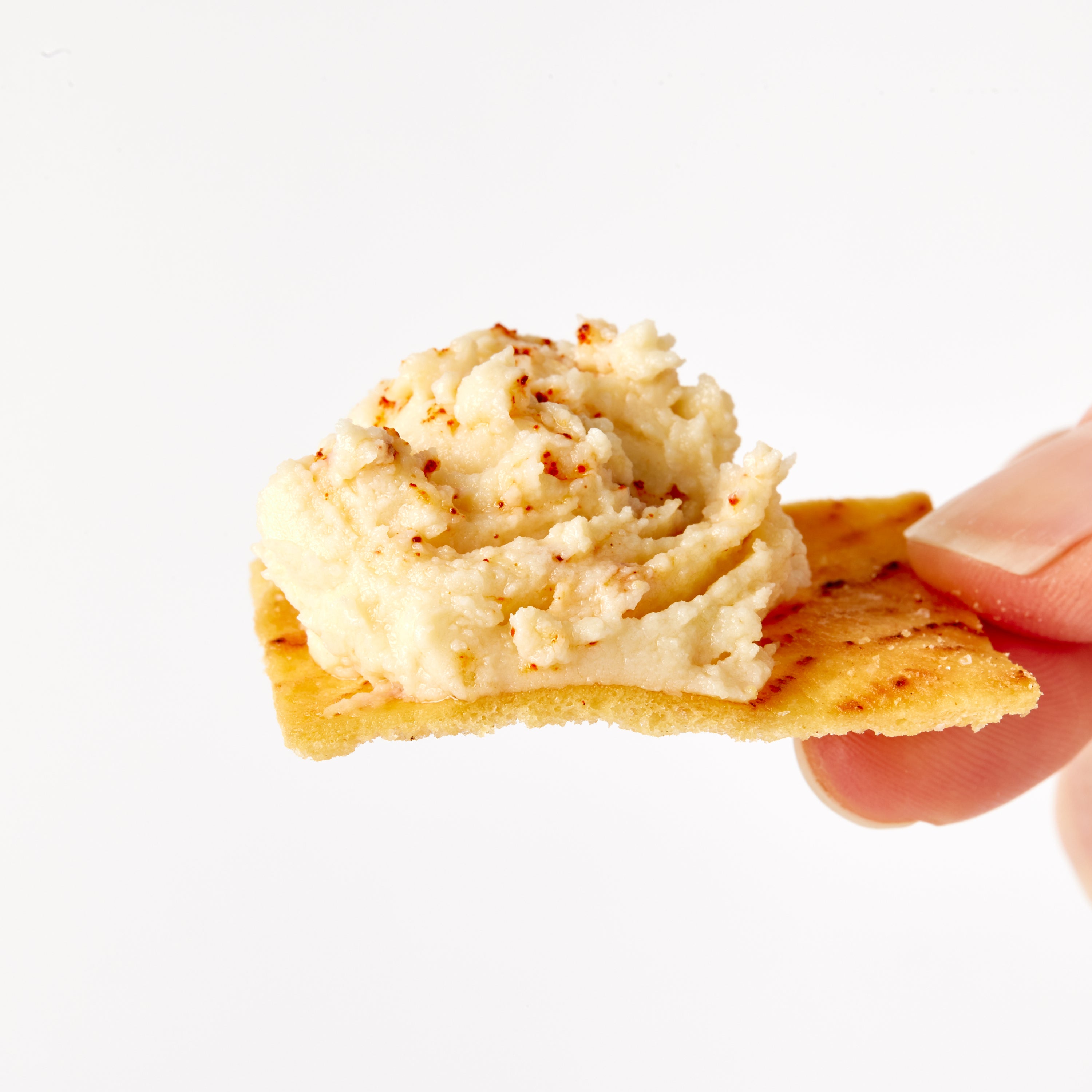 A close-up of a hand holding a square pita chip topped with a scoop of hummus with small red seasoning specks, against a plain white background.