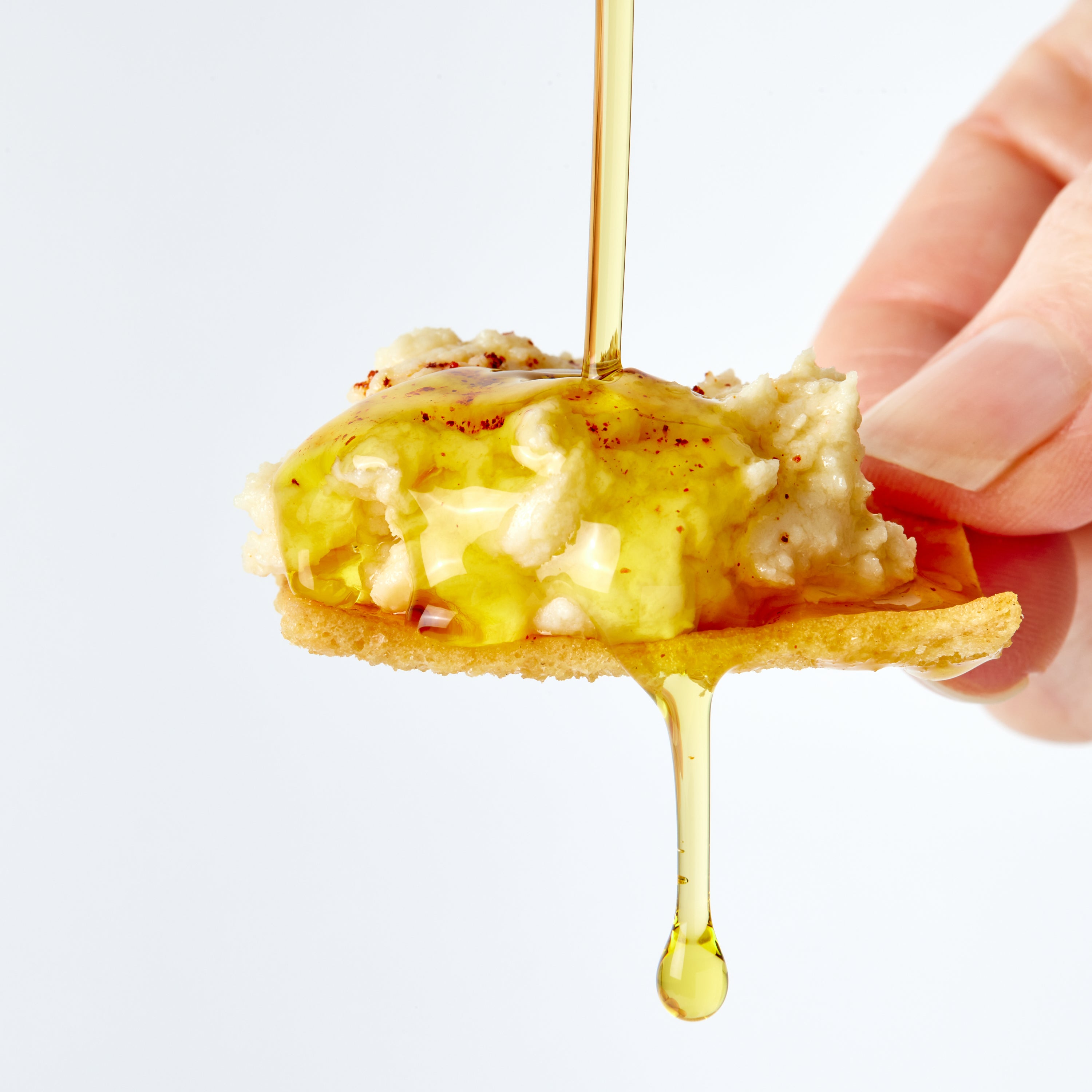A close-up of a hand holding a pita chip topped with hummus and drizzled with golden olive oil against a clean white background.