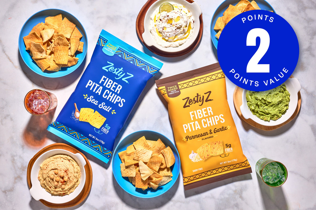 Two bags of ZestyZ Fiber Pita Chips arranged on a light marble surface with bowls of chips, dip, guacamole, and salsa around them. A large blue circular badge in the top right says '2 POINTS VALUE'.